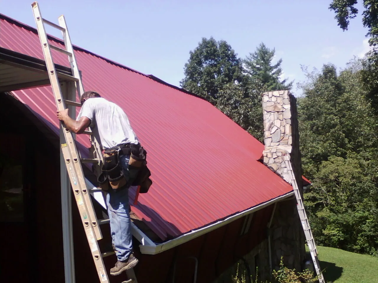 Roofer on a ladder beside a red metal roof and stone chimney — Soffit & Fascia Repair by Roofing Brothers in Suffern